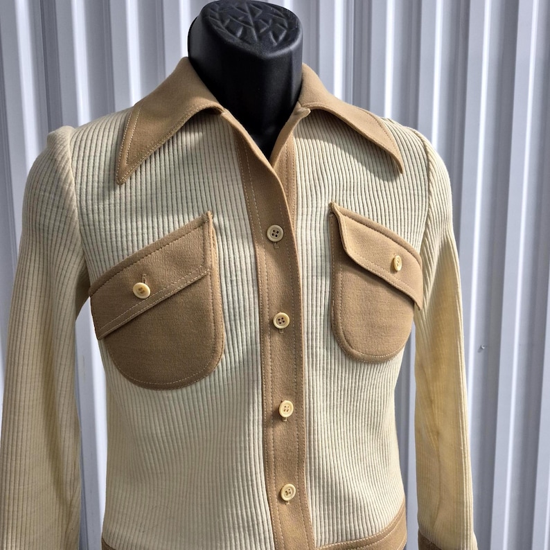 Cream-colored ribbed knit shirt with tan accents. The shirt features a wide tan collar, two front pockets, and tan trim along the button closure and cuffs. The shirt is buttoned up and displayed on a mannequin. Size 8, By Act III, a vintage company of Jonathan Logan.