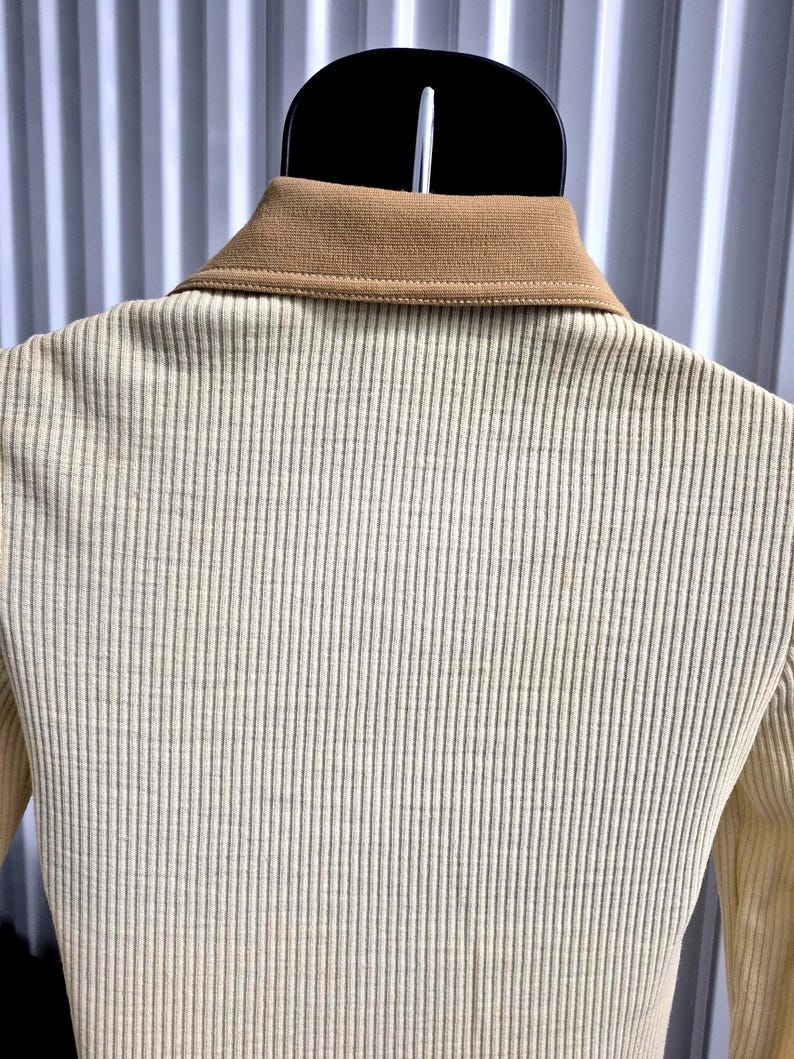 Another back view, Close-up of a cream-colored ribbed shirt with a tan collar. The shirt is hanging on a black hanger mannequin. The ribbed texture runs vertically down the shirt. The collar is a solid tan color.
