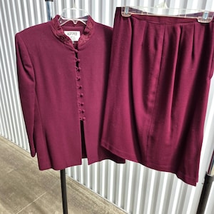 May include: A burgundy two-piece suit, consisting of a long-sleeved jacket with a high collar and button closure, and a matching skirt. The jacket has decorative embroidery around the collar. The suit is hanging on a clothing rack.