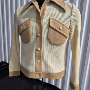 60s Cream-colored button-up jacket styled top with tan (caramel colored) accents. The jacket features a ribbed texture, a contrasting collar, two front pockets, and tan trim along the edges. The buttons are a matching cream color.