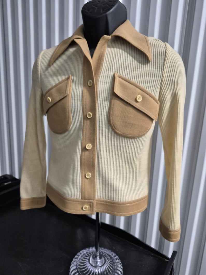60s Cream-colored button-up jacket styled top with tan (caramel colored) accents. The jacket features a ribbed texture, a contrasting collar, two front pockets, and tan trim along the edges. The buttons are a matching cream color.