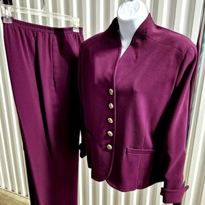 May include: A two-piece plum-colored suit displayed on a clothing rack. The suit includes a blazer with gold buttons and a pair of matching pants. The blazer has a notched collar and two front pockets. The pants have an elastic waistband.