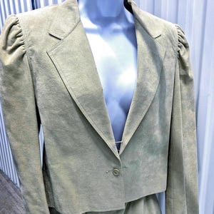 May include: A light olive green suede blazer featuring a single-button closure, notched lapels, and gathered puff shoulders. The jacket has a cropped silhouette and long sleeves, displayed on a mannequin against a vertical white background.