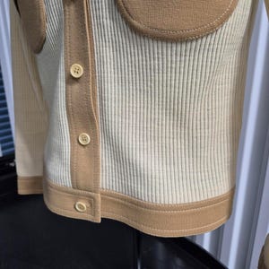 Beautiful Cream-colored ribbed knit cardigan with tan accents. Features contrasting button-down front detail, two patch pockets, and tan trim along the edges. Displayed on a mannequin, showcasing its tailored fit and design.