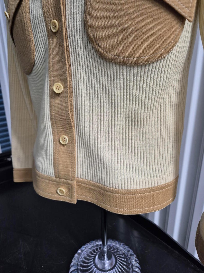 Beautiful Cream-colored ribbed knit cardigan with tan accents. Features contrasting button-down front detail, two patch pockets, and tan trim along the edges. Displayed on a mannequin, showcasing its tailored fit and design.