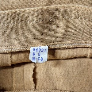 Close-up of the waistband of this vintage 60s tan and cream 2 piece skirt set. A small, rectangular tag is attached, displaying the numbers "#6031" and "M-S8" for Misses size 8. The fabric appears to be a knit or woven material, with visible seams and stitching.