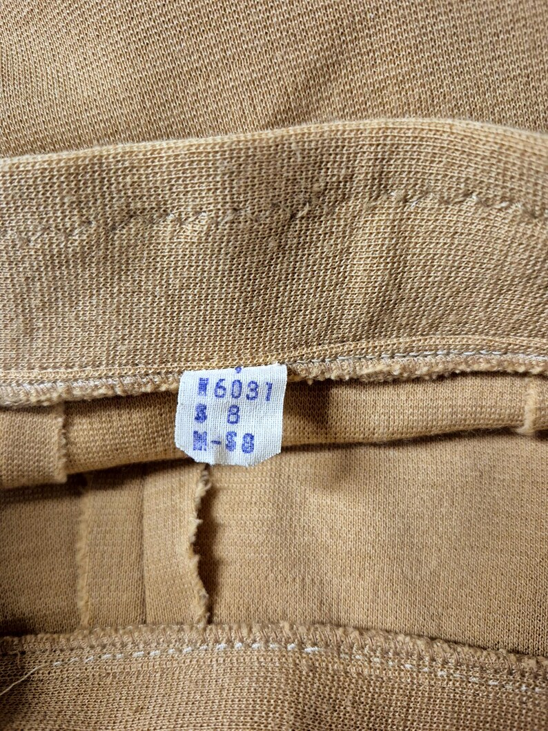 Close-up of the waistband of this vintage 60s tan and cream 2 piece skirt set. A small, rectangular tag is attached, displaying the numbers "#6031" and "M-S8" for Misses size 8. The fabric appears to be a knit or woven material, with visible seams and stitching.