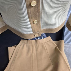 A two-piece caramel and cream-colored knitwear set. The top is a button-down jacket style cardigan with ribbed detailing and tan trim. The matching skirt is a solid tan color with a flared design. The set is made of a soft, comfortable material.