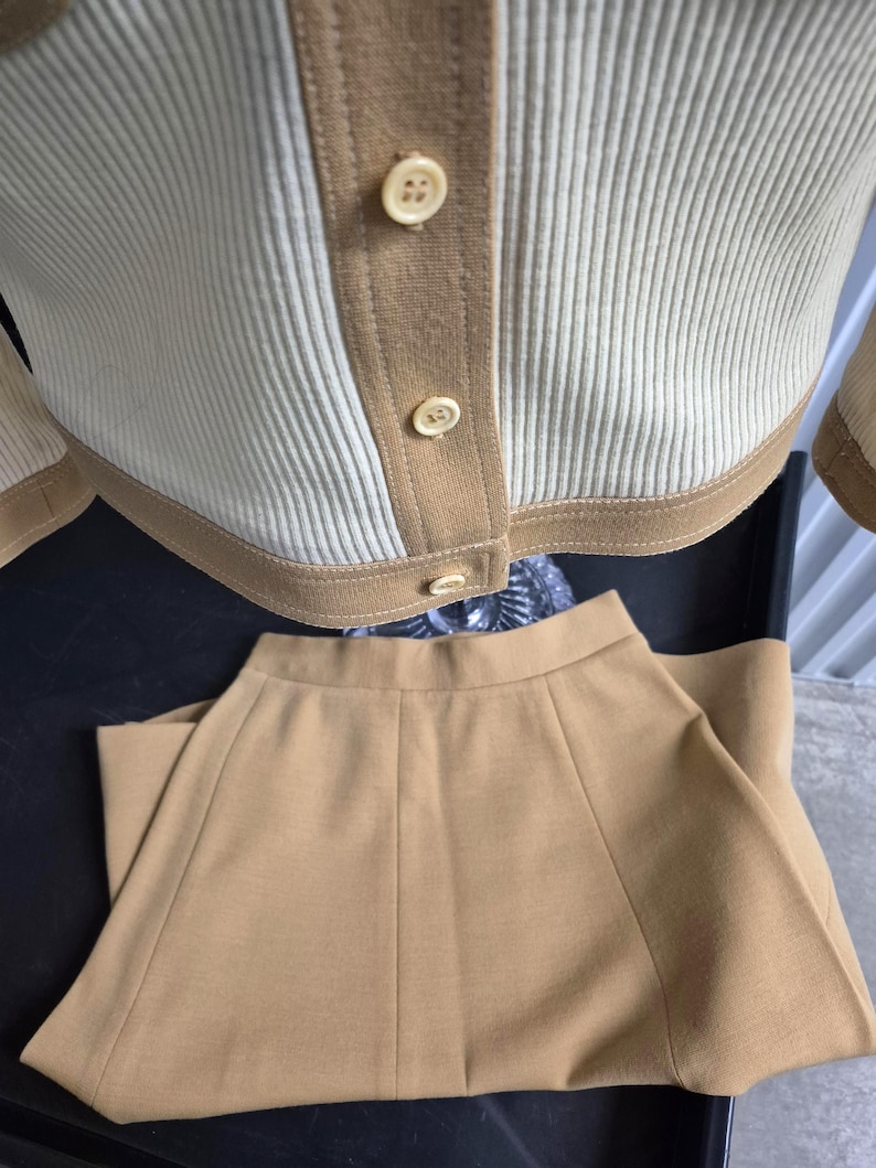 A two-piece caramel and cream-colored knitwear set. The top is a button-down jacket style cardigan with ribbed detailing and tan trim. The matching skirt is a solid tan color with a flared design. The set is made of a soft, comfortable material.