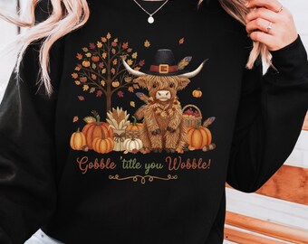 May include: Black sweatshirt featuring a whimsical illustration of a Highland cow wearing a pilgrim hat, surrounded by pumpkins, a tree with autumn leaves, and the text "Gobble 'title you Wobble!"