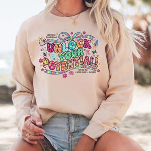 Könnte beinhalten: Beiges Sweatshirt mit dem Text "UNLOCK YOUR POTENTIAL!" in farbenfrohen Buchstaben im Retro-Stil. Das Design umfasst eine Schleife, Blumen und den Satz "DREAM BIG + WORK HARD = SUCCESS."