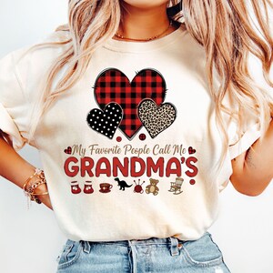 May include: Cream-colored t-shirt featuring a graphic with three hearts: a red and black buffalo plaid heart, a black polka dot heart, and a leopard print heart. The text reads "My Favorite People Call Me GRANDMA'S" with small illustrations below.