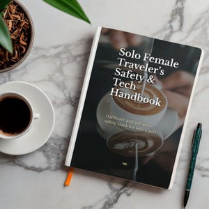 Può includere: Un libro intitolato "Solo Female Traveler's Safety & Tech Handbook" con un'immagine di tazza di caffè sulla copertina. Una tazza di caffè su un piattino, una penna e una piccola pianta sono su una superficie di marmo.