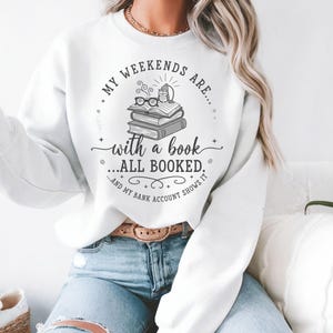 May include: White sweatshirt featuring the text "MY WEEKENDS ARE... with a book... ALL BOOKED... AND MY BANK ACCOUNT SHOWS IT" in black. The design includes a stack of books, glasses, and a small light.