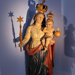 Könnte beinhalten: Geschnitzte Holzstatue der Jungfrau Maria, die ein Kind hält, beide gekrönt. Die Statue ist in gedämpften Tönen von Creme, Rot und Schwarz bemalt. Die Jungfrau Maria hält ein sternförmiges Zepter. Die Statue steht auf einem schwarzen Sockel.