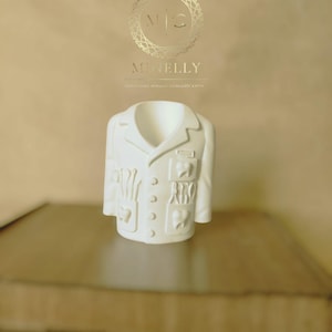 Könnte beinhalten: Eine weiße Keramikskulptur eines Arztkittels mit Taschen, Knöpfen und Revers. Die Skulptur steht auf einer braunen Oberfläche. Das goldene Logo von "MINELLY" ist ebenfalls zu sehen.