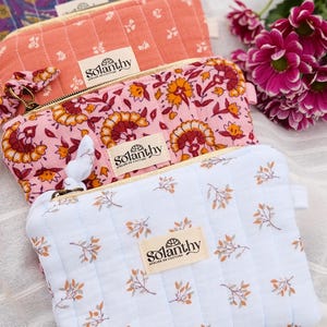 Peut inclure: Gros plan sur quatre trousses de maquillage matelassées de différentes couleurs et motifs. Les sacs présentent des motifs floraux et géométriques dans des tons de rose, d'orange et de bleu. Chaque sac a une fermeture éclair et une étiquette "Solanthy Atelier de Couture."