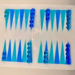 May include: A backgammon board with a white surface, featuring alternating blue and teal triangles. Translucent blue game pieces are arranged on the board, ready for play. The board is a classic game for two players.