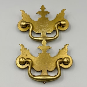 May include: Two antique brass drawer pulls. Each pull has an ornate design with a curved handle and two circular mounting points. The brass has a slightly aged, tarnished appearance, suggesting vintage use.