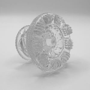 May include: Clear glass decorative object with a detailed, textured design. The object has a circular base with a floral pattern and a smaller, cylindrical stem. The glass is transparent, allowing light to pass through.