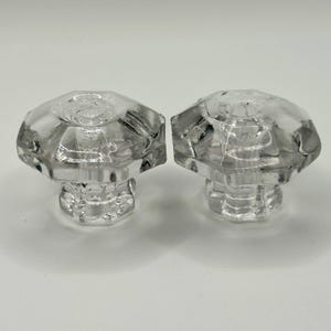 May include: Two clear, faceted glass knobs with a hexagonal top and a cylindrical base. The knobs are likely for a cabinet or drawer, and are set against a white background.