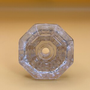 Könnte beinhalten: Klarer, achteckiger Glasflaschenverschluss mit einem zentralen Loch. Der Verschluss hat ein detailliertes, strukturiertes Design und besteht aus transparentem Glas. Das Design ist symmetrisch und geometrisch.