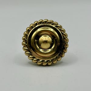 Könnte beinhalten: Ein runder, goldfarbener Knopf mit einem zentralen kreisförmigen Design und einem seilartigen Rand. Der Knopf ist aus Metall und hat eine glänzende Oberfläche. Die Hardware ist wahrscheinlich für einen Schrank oder eine Schublade. Durchmesser: ca. 3 cm.