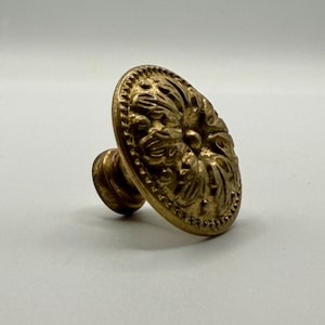 May include: A brass-colored cabinet knob with an oval, ornate face featuring a floral design. The knob has a short, cylindrical stem and a detailed, textured surface. The overall design is vintage.