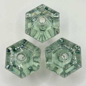 Puede incluir: Tres tiradores hexagonales de cristal en tono verde pálido. Cada tirador presenta un orificio central para el montaje y bordes facetados, creando una estética vintage. Los tiradores están dispuestos en un patrón triangular sobre un fondo blanco.