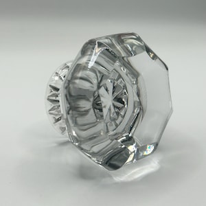 Könnte beinhalten: Klarer, facettierter Türknauf aus Glas mit achteckiger Form. Der Knauf hat eine dekorative, kreisförmige Basis mit einem Sternmuster in der Mitte. Das Glas reflektiert das Licht und erzeugt einen funkelnden Effekt.
