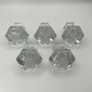 May include: Five clear glass cabinet knobs with a faceted, hexagonal design. The knobs are transparent, allowing light to pass through, and are arranged against a white background. These knobs are suitable for furniture restoration or new builds.