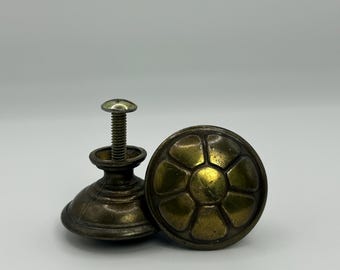 May include: Two antique brass cabinet knobs. One knob is round with a floral design, and the other has a screw attached. The knobs have a tarnished, aged appearance, suggesting they are vintage or vintage-inspired.