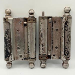 Puede incluir: Dos bisagras metálicas antiguas con un acabado envejecido. Cada bisagra tiene un cuerpo cilíndrico con remates decorativos en forma de bola en la parte superior e inferior. El metal muestra signos de desgaste, con zonas de pintura negra y óxido.