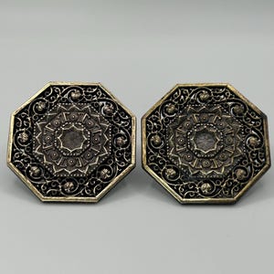 May include: Two ornate, octagonal metal objects with intricate filigree and a central circular design. The objects have a dark, antique finish with gold-colored edges, suggesting a vintage or decorative item.