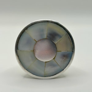 May include: A round, silver-toned cabinet knob with a mother-of-pearl inlay. The inlay is composed of iridescent, segmented pieces arranged in a circular pattern around a central, pinkish-silver disc. The knob has a simple, elegant design.