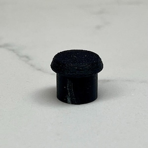 May include: A small, black, cylindrical object with a rounded, textured top. The object appears to be made of a solid material and is set against a light-colored background. The design is simple and minimalist.