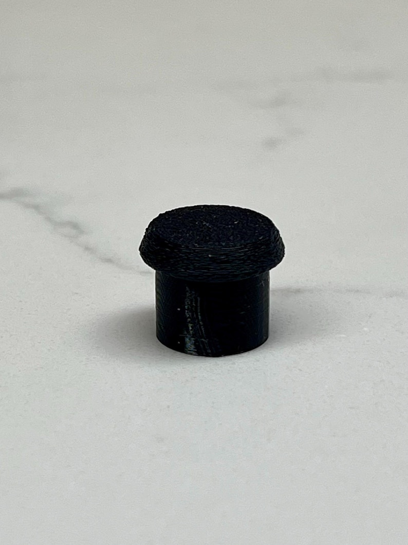 May include: A small, black, cylindrical object with a rounded, textured top. The object appears to be made of a solid material and is set against a light-colored background. The design is simple and minimalist.