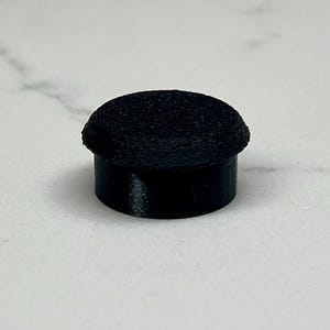 May include: A black, cylindrical object with a textured, rounded top. The object has a short, smooth base and a slightly wider, textured upper portion. The item is set against a light-colored background.