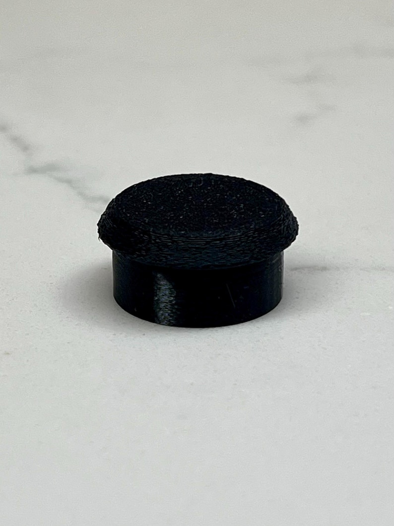 May include: A black, cylindrical object with a textured, rounded top. The object has a short, smooth base and a slightly wider, textured upper portion. The item is set against a light-colored background.