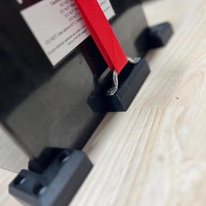 Puede incluir: Objeto rectangular negro con una correa roja y el texto "FPV-P". El objeto tiene soportes de plástico negro y una etiqueta blanca con texto. La correa está asegurada con herrajes metálicos.