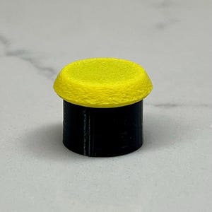 May include: A small, cylindrical object with a bright yellow top and a black base. The top has a textured surface, and the base appears smooth. The object is set against a light gray background.