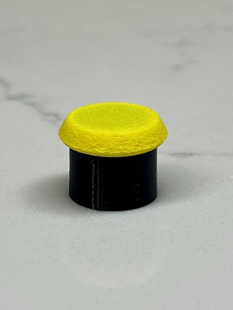 May include: A small, cylindrical object with a bright yellow top and a black base. The top has a textured surface, and the base appears smooth. The object is set against a light gray background.