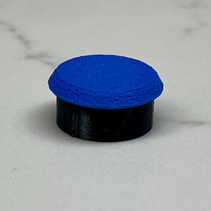 May include: A small, cylindrical object with a blue, textured top and a black base. The top appears to be made of layered material. The object is set against a white background.