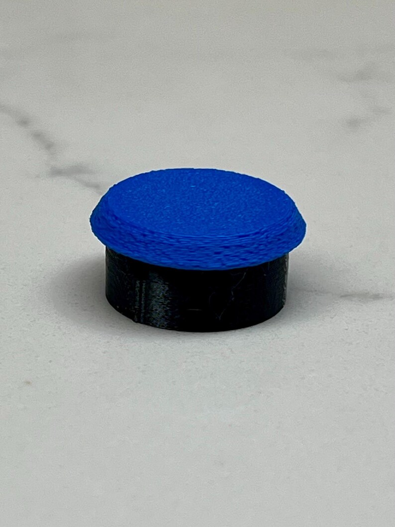 May include: A small, cylindrical object with a blue, textured top and a black base. The top appears to be made of layered material. The object is set against a white background.