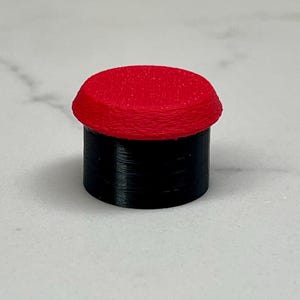 May include: A small, cylindrical object with a red, rounded top and a black base. The top has a textured surface. The object is likely a button or a small decorative piece.