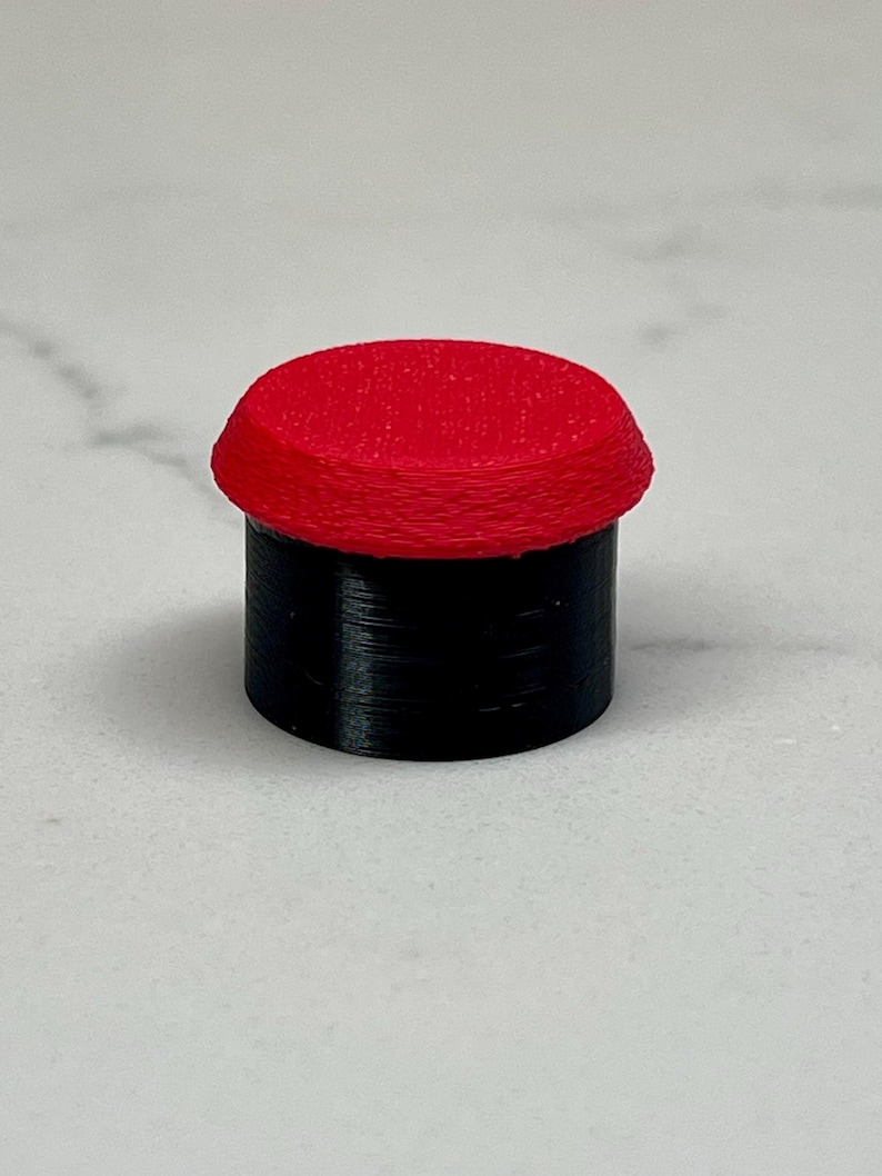 May include: A small, cylindrical object with a red, rounded top and a black base. The top has a textured surface. The object is likely a button or a small decorative piece.