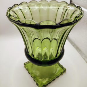 May include: A green glass vase with a scalloped rim and a flared, ribbed body. The vase sits on a square, decorative base. The glass has a slight transparency, allowing light to pass through. The vase is a vintage piece.