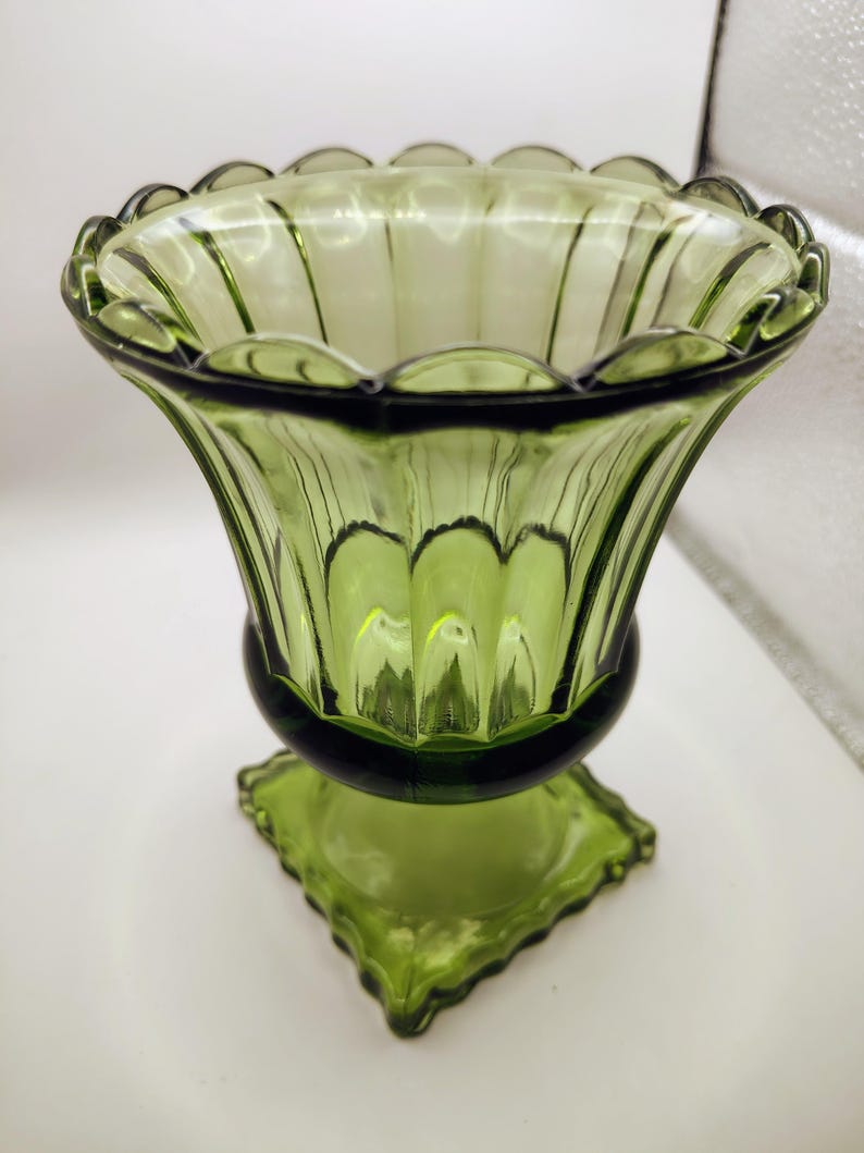 May include: A green glass vase with a scalloped rim and a flared, ribbed body. The vase sits on a square, decorative base. The glass has a slight transparency, allowing light to pass through. The vase is a vintage piece.