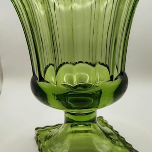 May include: A green glass candle holder with a fluted, tulip-shaped bowl and a square base. The glass has a ribbed texture and a scalloped rim. The candle holder is a decorative item.