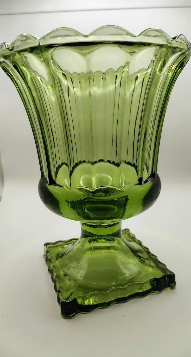 May include: A green glass candle holder with a fluted, tulip-shaped bowl and a square base. The glass has a ribbed texture and a scalloped rim. The candle holder is a decorative item.